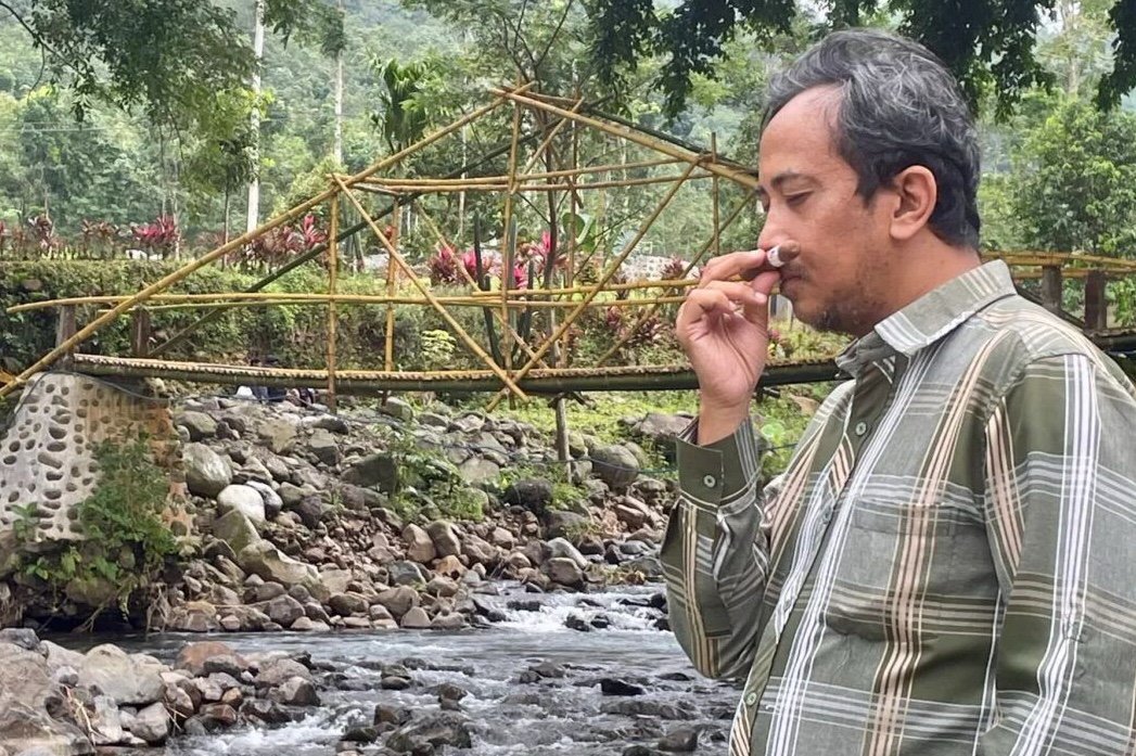 Man by river, bamboo bridge, touching nose.