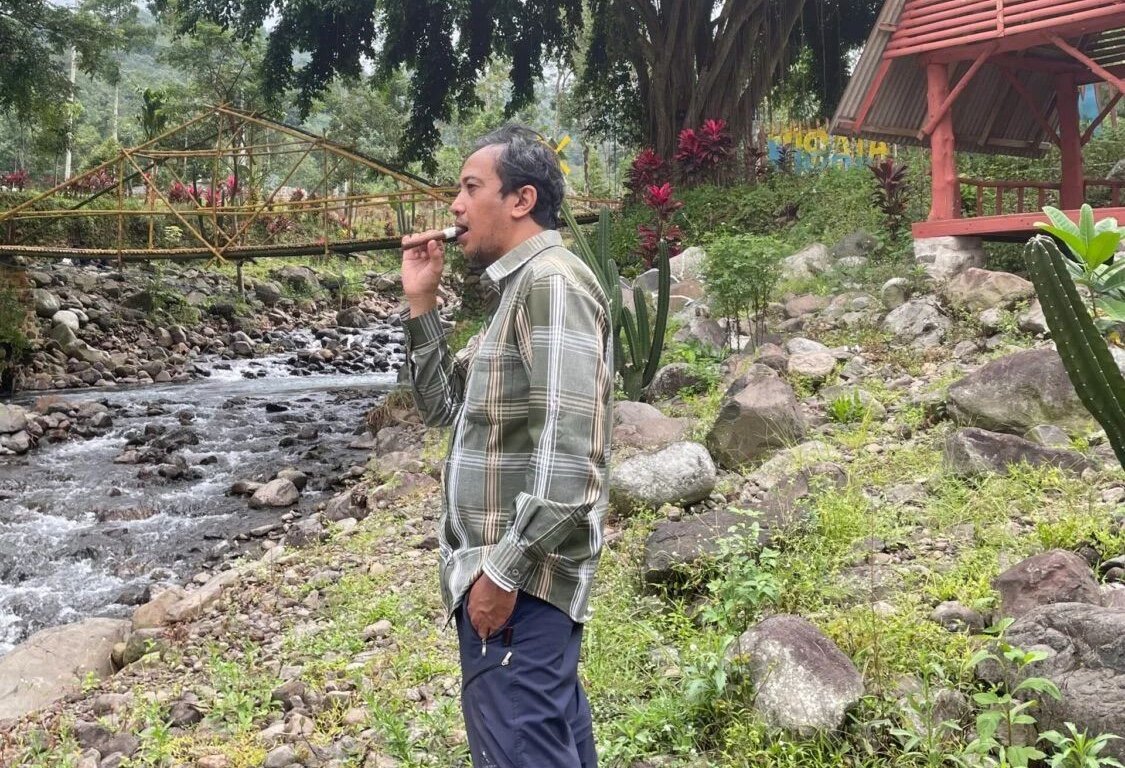 Man smoking cigar by a rocky river with a bamboo bridge.