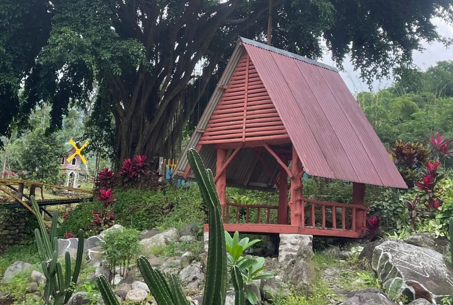 Red gazebo, large tree, cacti, and windmill in a garden.
