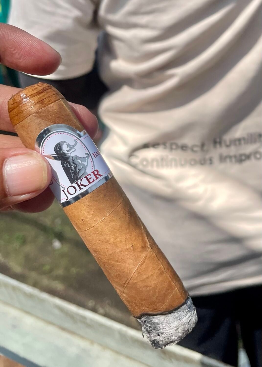 Hand holding a lit Joker brand cigar with ash.